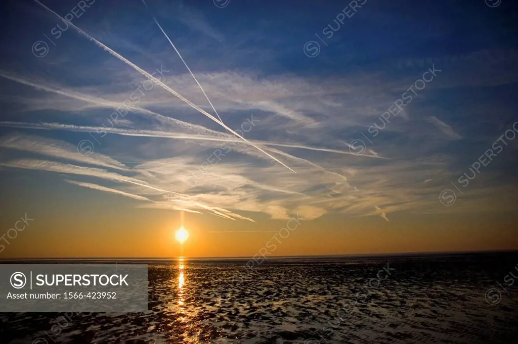 A setting sun creates an orange layer in the deep blue sky and blazes a gold trail across the wet sand of the beach. White vapour trails criss-cross t...