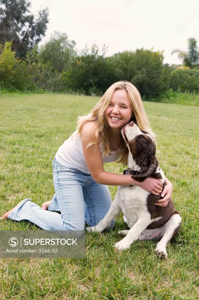 Smiling blonde teen girl in blue jeans and white tank top kneeling on grass lawn, get´s a kiss from her pet dog.