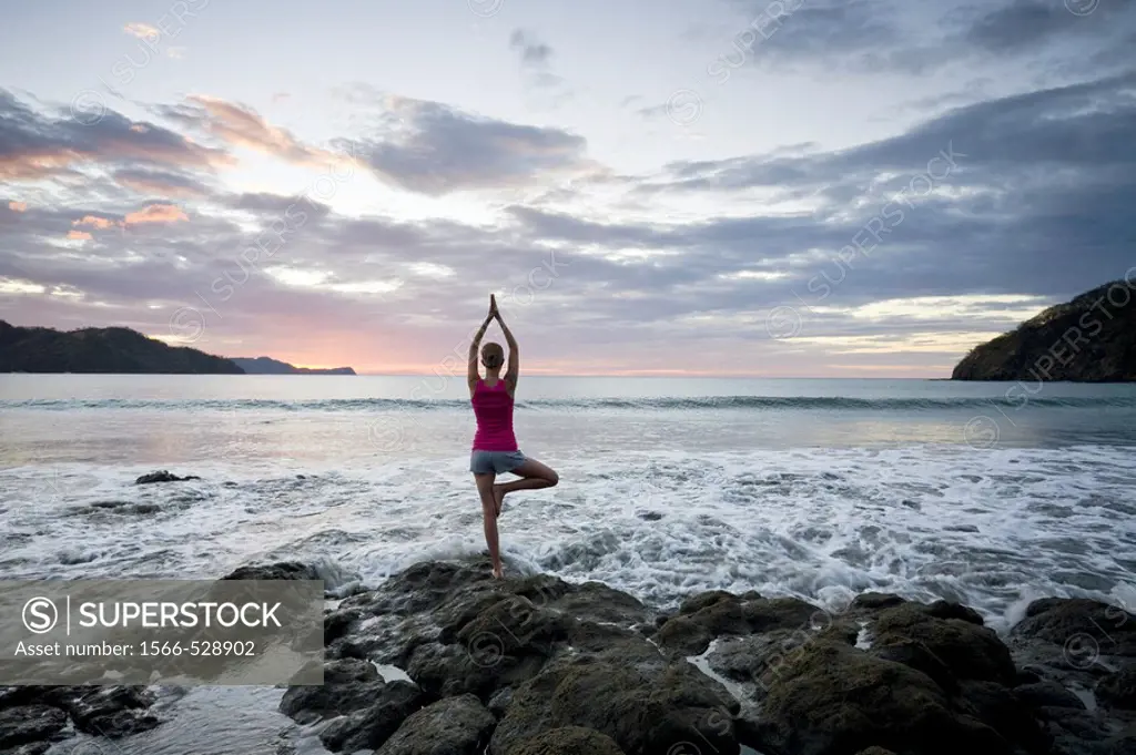Young woman doing yoga on rocks at sunset in front of ocean surf at Playas del Coco, Costa Rica