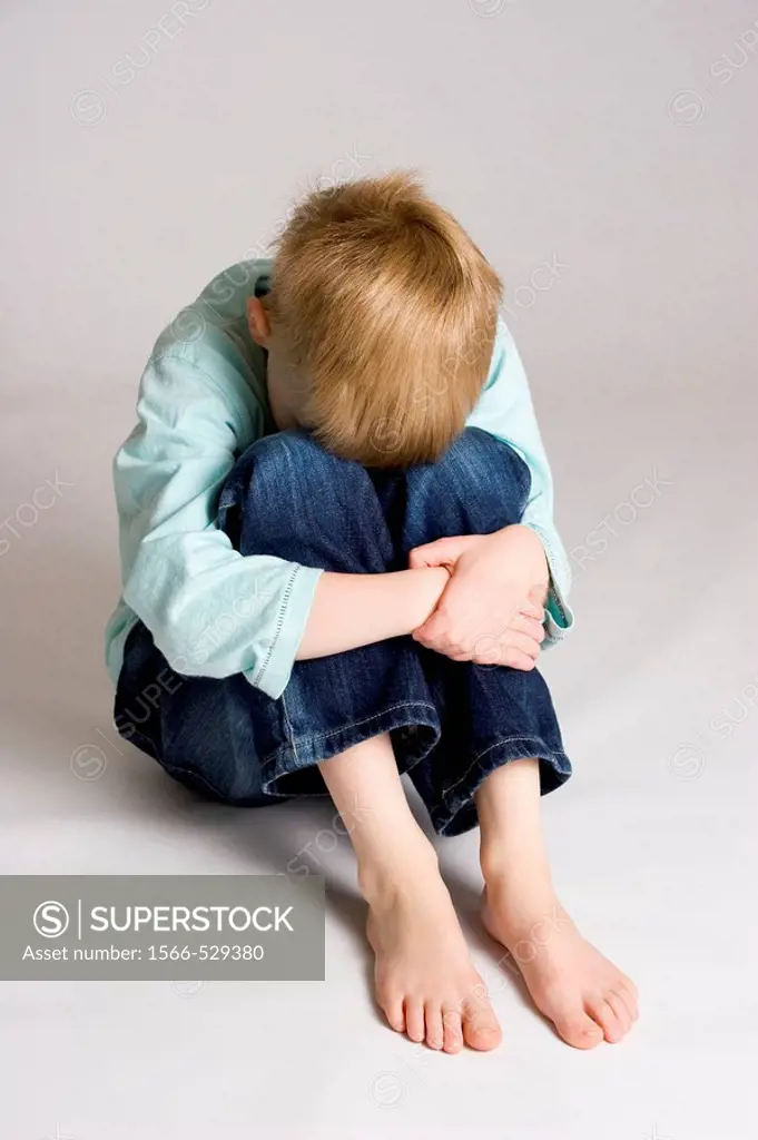 A young boy sits hunched over hugging his bent legs and hiding his face