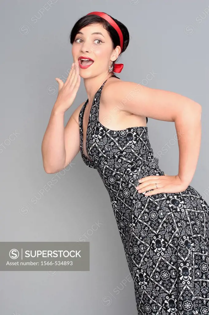 A 20-something attractive female in a sleeveless dress with a red ribbon in her hair has one hand on her hip and the other held to her mouth expressin...