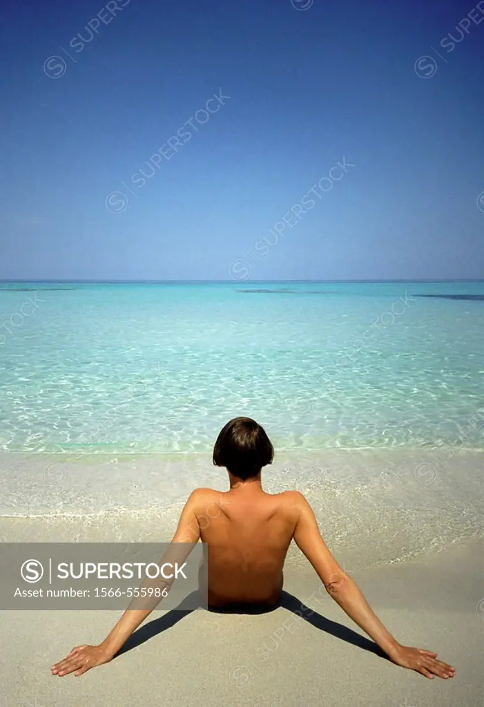Naked woman sitting on sand at the beach looking out to sea