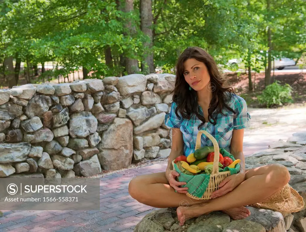 32 year old brunette woman in casual summer dress sitting cross-legged on a stone wall holding a basket of mixed vegetables.
