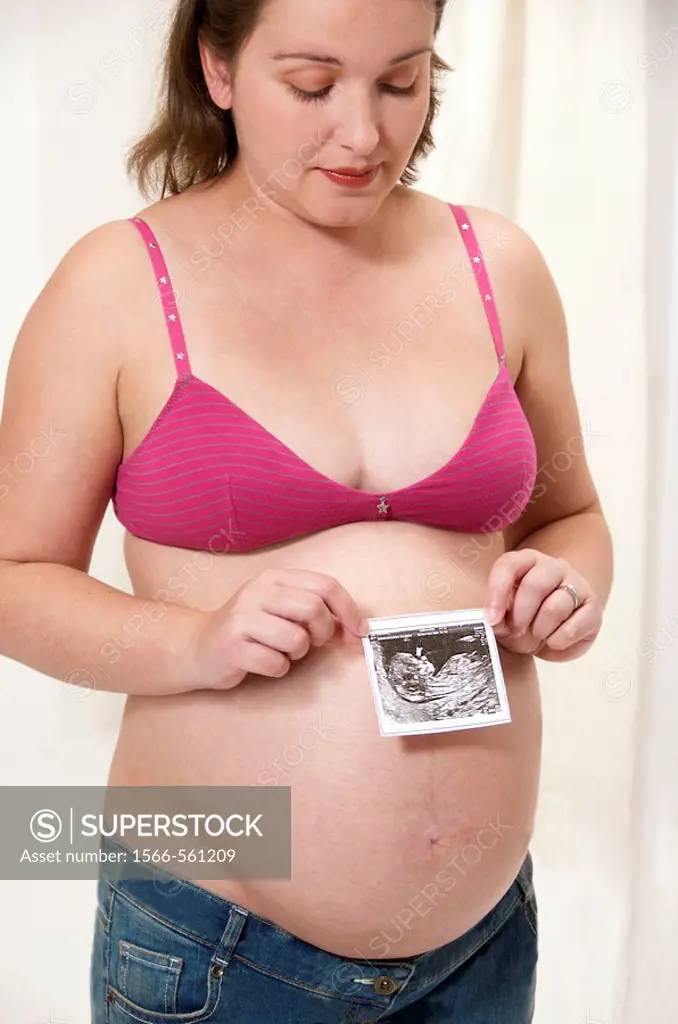 Young pregant woman in a pink brassiere and jeans, with her protruding belly, holding the echography of her future baby