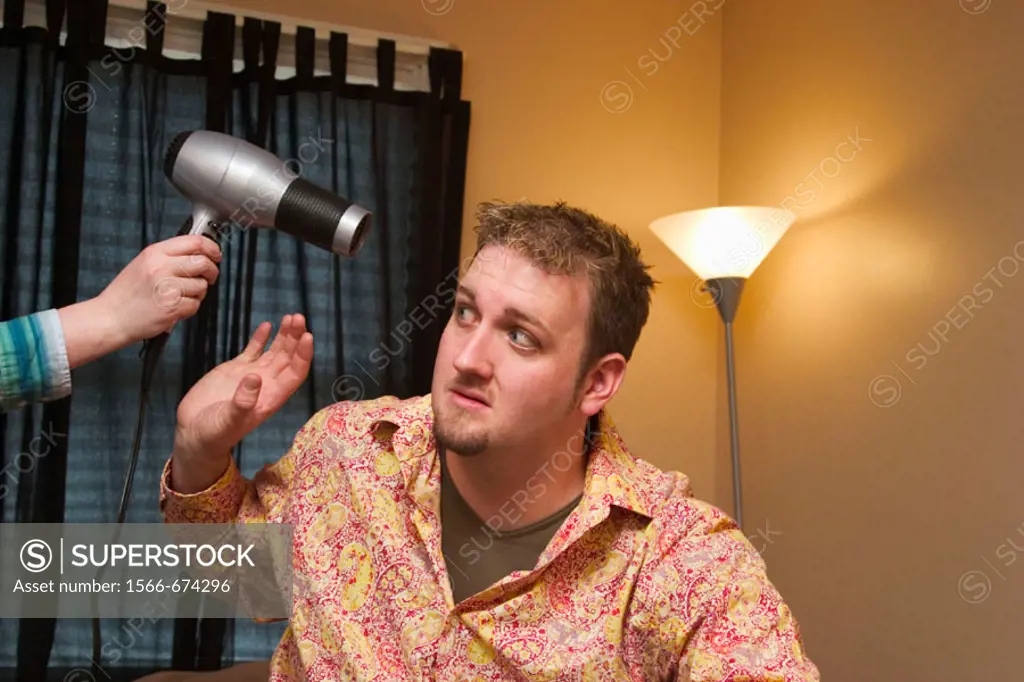 Young man sitting on the bed in his bedroom, with a young woman´s hand holding a hair dryer by his head.