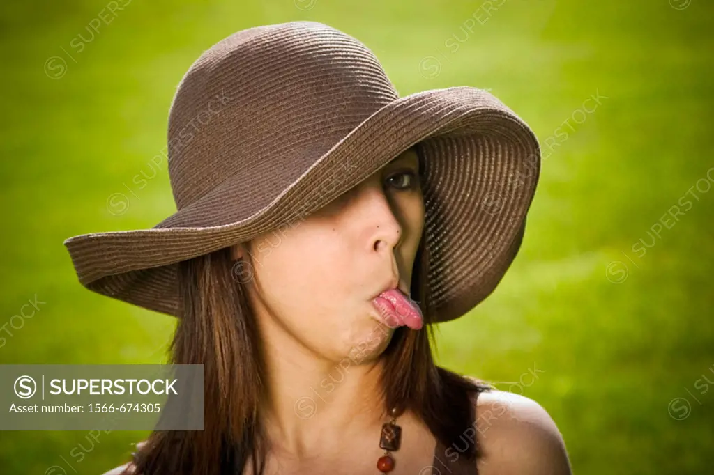 Young Spanish/Asian woman, wearing a hat, sticking out her tongue while posing at a park.