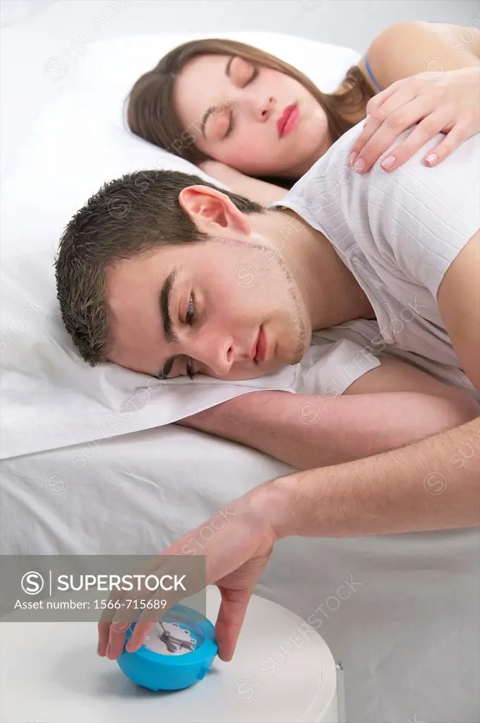 Young couple in bed, he is awake and turning off the alarm clock, she is fast asleep, and holding him, tenderly