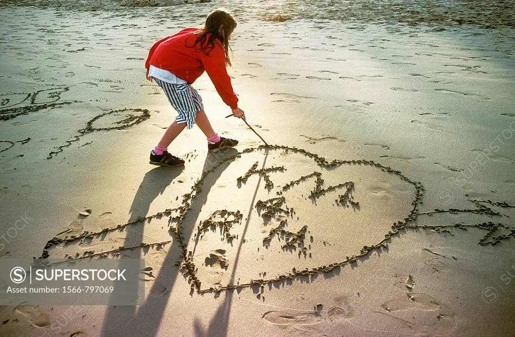 Young girl draws a love heart in the sand at the beach as the sun fades into an evening glow