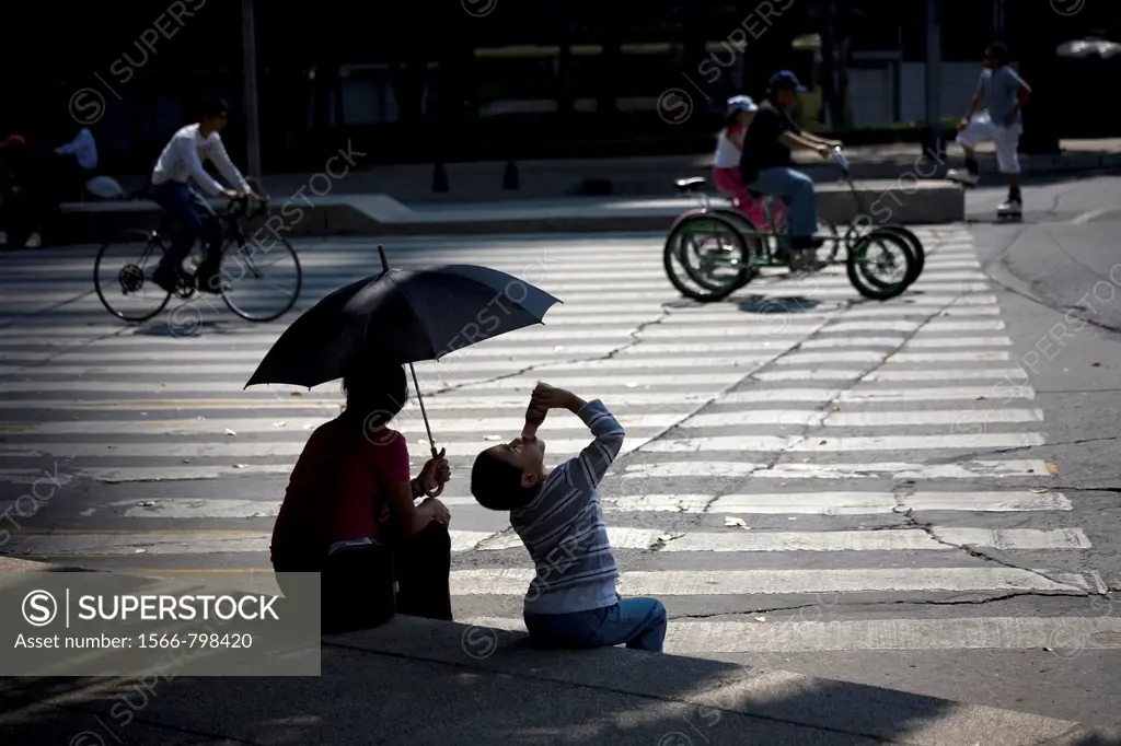 A boy drinks yogurt next to his mother holding an umbrella to shade them from the sun as they watch cyclists ride along Paseo de la Reforma Avenue in ...