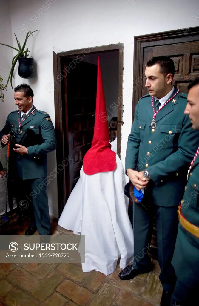 A penitent wearing a red pointed hood and a white cape pass by Guardia Civil members, a Spanish military-status police force inside a church as he wai...