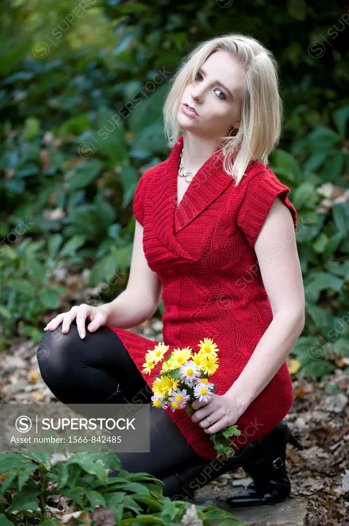 21 year old blond woman wearing a red sweater and black boots and stockings holding a bouquet of yellow flowers on a garden path looking at camera.