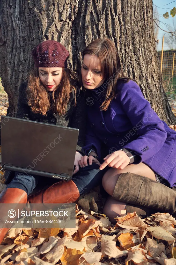 Two young women outdoors sitting on autumn leaves using laptop