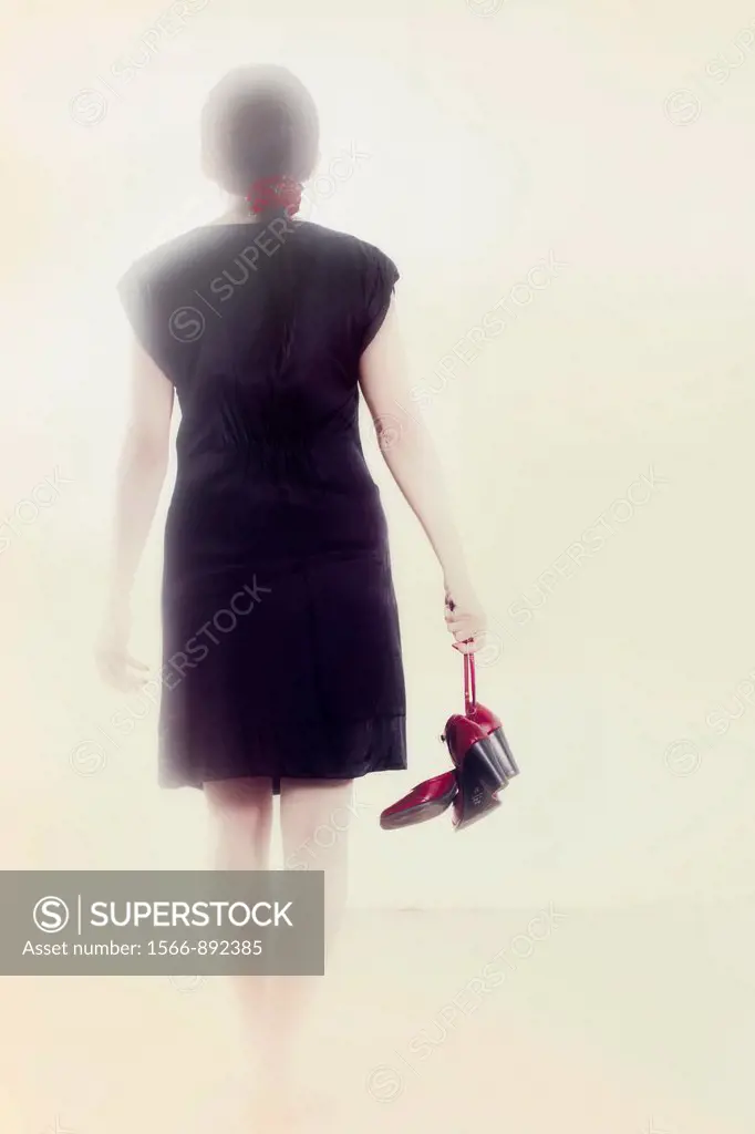 Woman in a black dress goes with red shoes in hand into the light