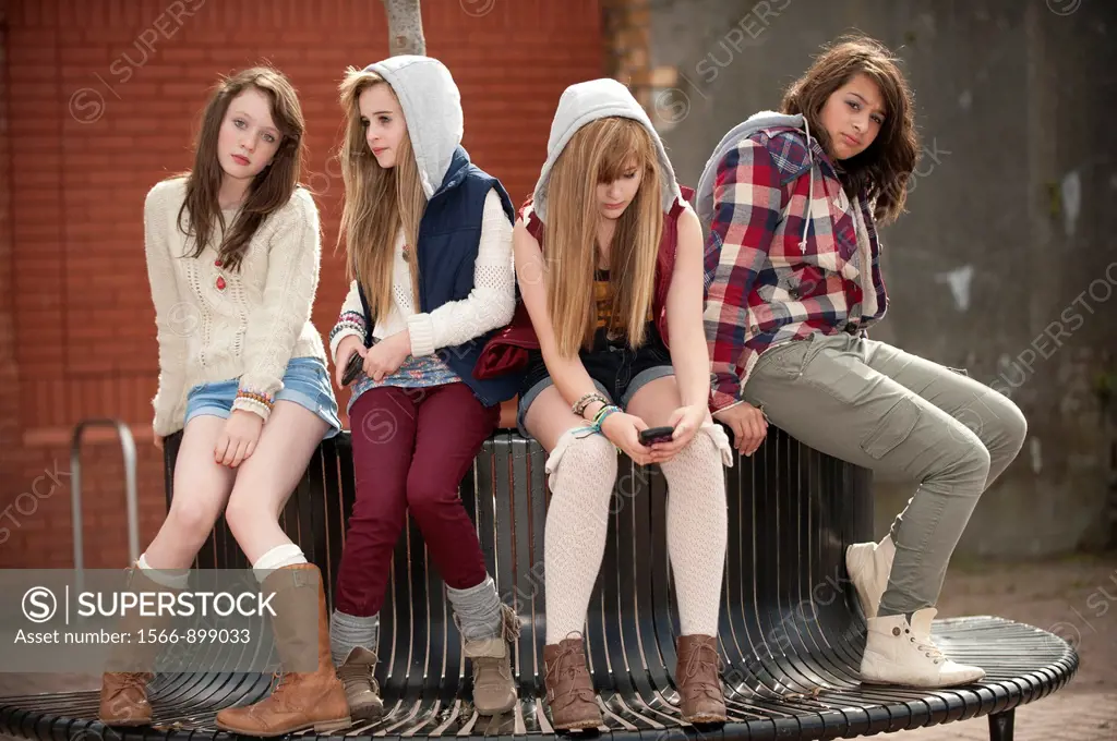 A group of 13 year old teenage girls, looking bored moody with attitude, sitting on a bench UK