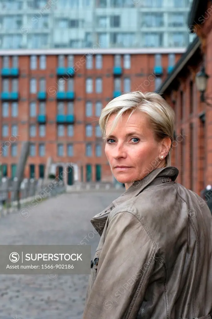 Blond haired executive woman, turning her head and looking back over her shoulder, seen from backside, with office building made of red bricks, window...