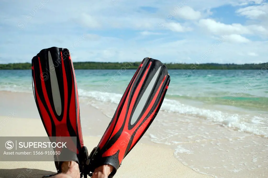 Close up of two red fins with male feet, holding up the fins at the beach with the turquise sea and blue sky with clouds in the background, sunny day,...