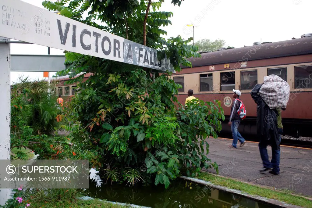 Close-up of the station sign ´Victoria Falls´ station with the platform, people passing by and carrying their luggage, a local passenger train in the ...