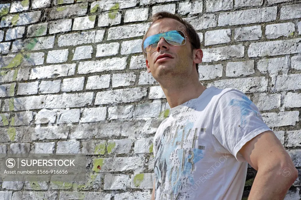Half body image of young caucasian man, looking cool with sunglasses into the far distance, T-Shirt, seen from front, wall of old white bricks behind ...