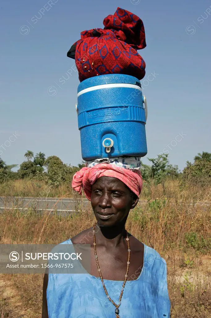 Woman carries belongings atop liquid dispenser on head near Kartong The Gambia