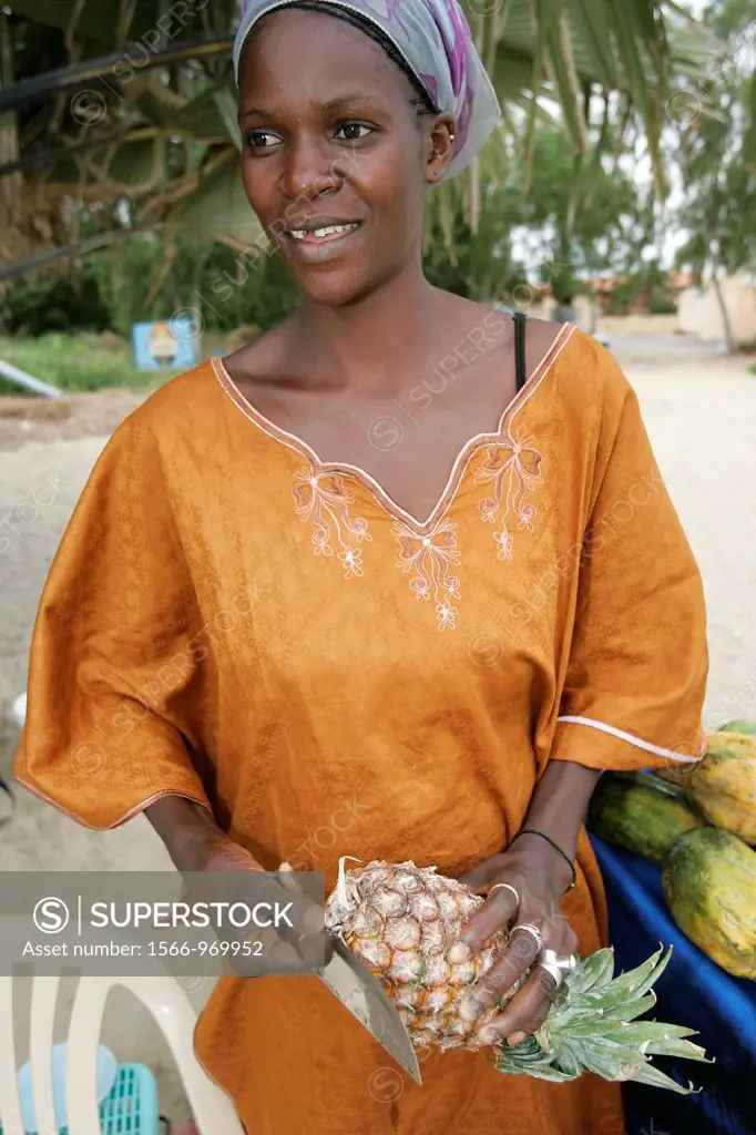 Woman fruit vendor in traditional dress peels pineapple The Cape beach Bakau The Gambia