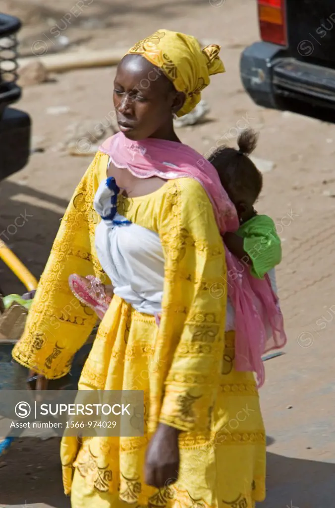 Baby in sling on back of woman walking in yellow traditional clothes Serekunda Market The Gambia