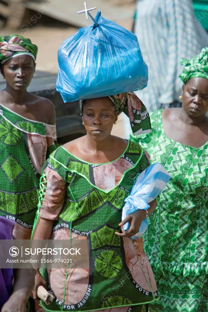 Woman in green traditional clothes carries plastic wrap bundle on head Serekunda Market The Gambia