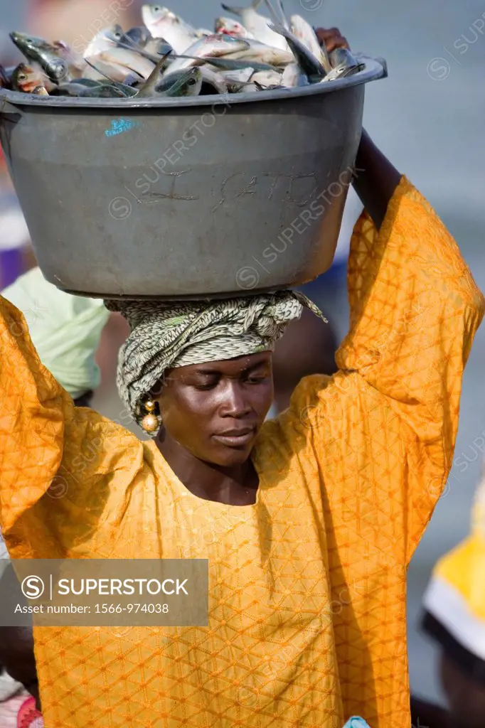 Young women carry basins of fresh fish on head up beach to market as open fishing boats arrive with catch Tanji The Gambia