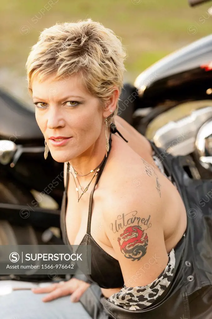 A provocative portrait of a 41 year old blond woman smiling at the camera wearing a leather vest and showing a tattoo on her left shoulder, motorcycle...