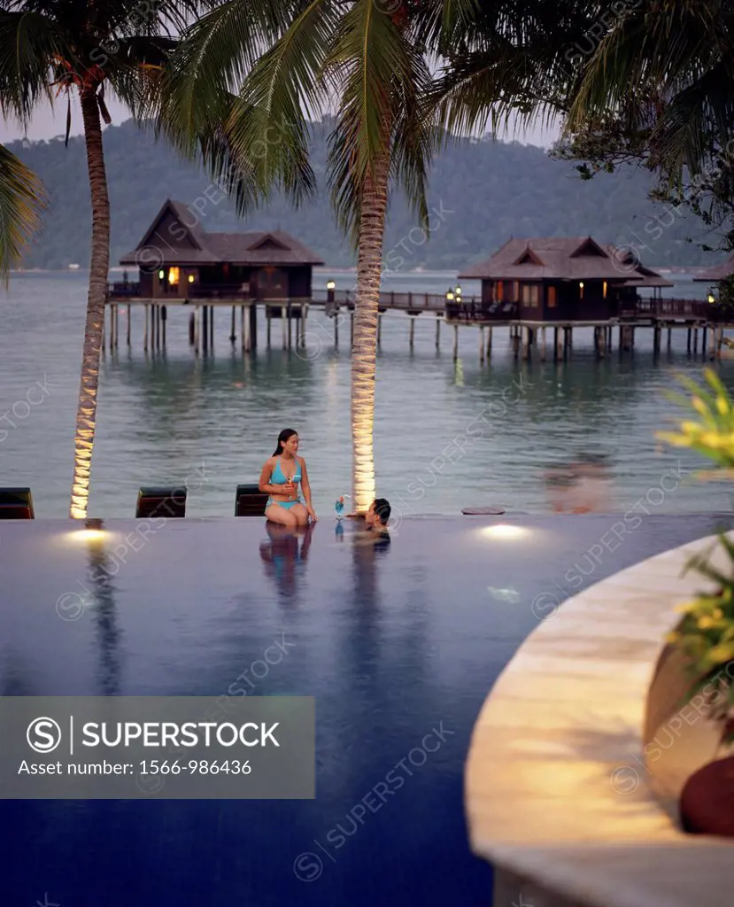 A woman and man sit on the edge of an infinity pool, sipping cocktails, at the Pangkor Laut Resort island in Malaysia, a famous resort known for it´s ...