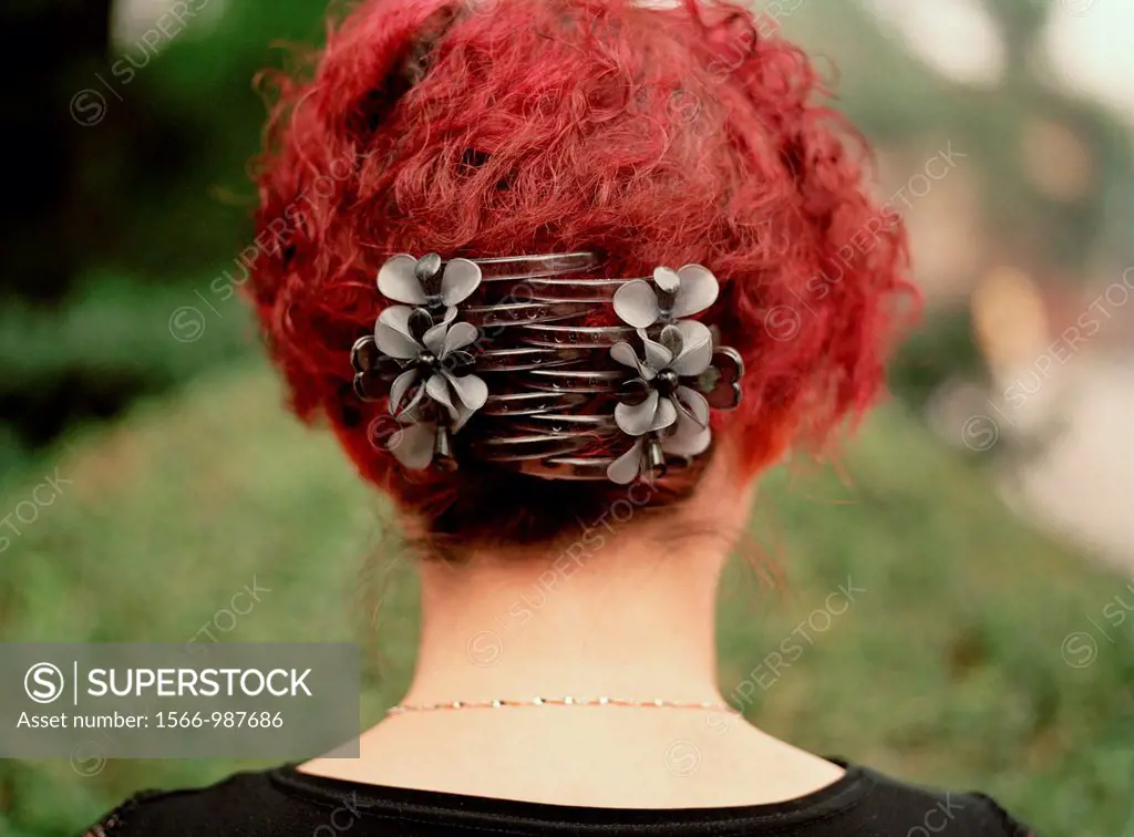 A portrait of the back of a Chinese woman´s head with bright red hair and large clips holding it in place, in a park
