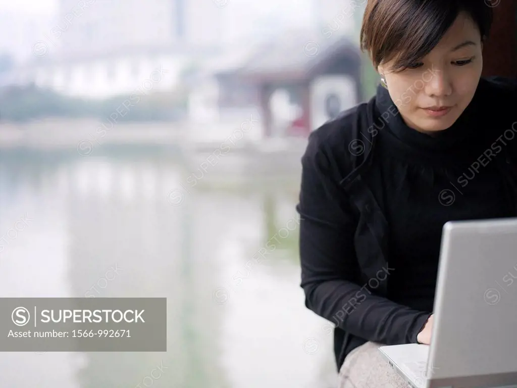 A Chinese business woman in a suit checking her email on her laptop through wireless internet with a lake and traditional Chinese architecture in the ...