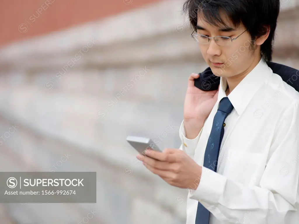 A young Chinese businessman in a shirt with a suit over his shoulder checking his text messages or email on his mobile smart phone through wireless in...