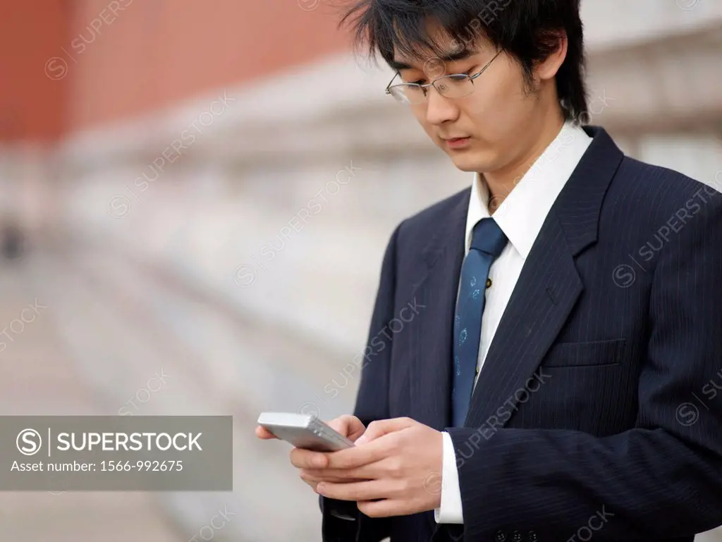 A young Chinese businessman in a shirt with a suit over his shoulder checking his text messages or email on his mobile smart phone through wireless in...