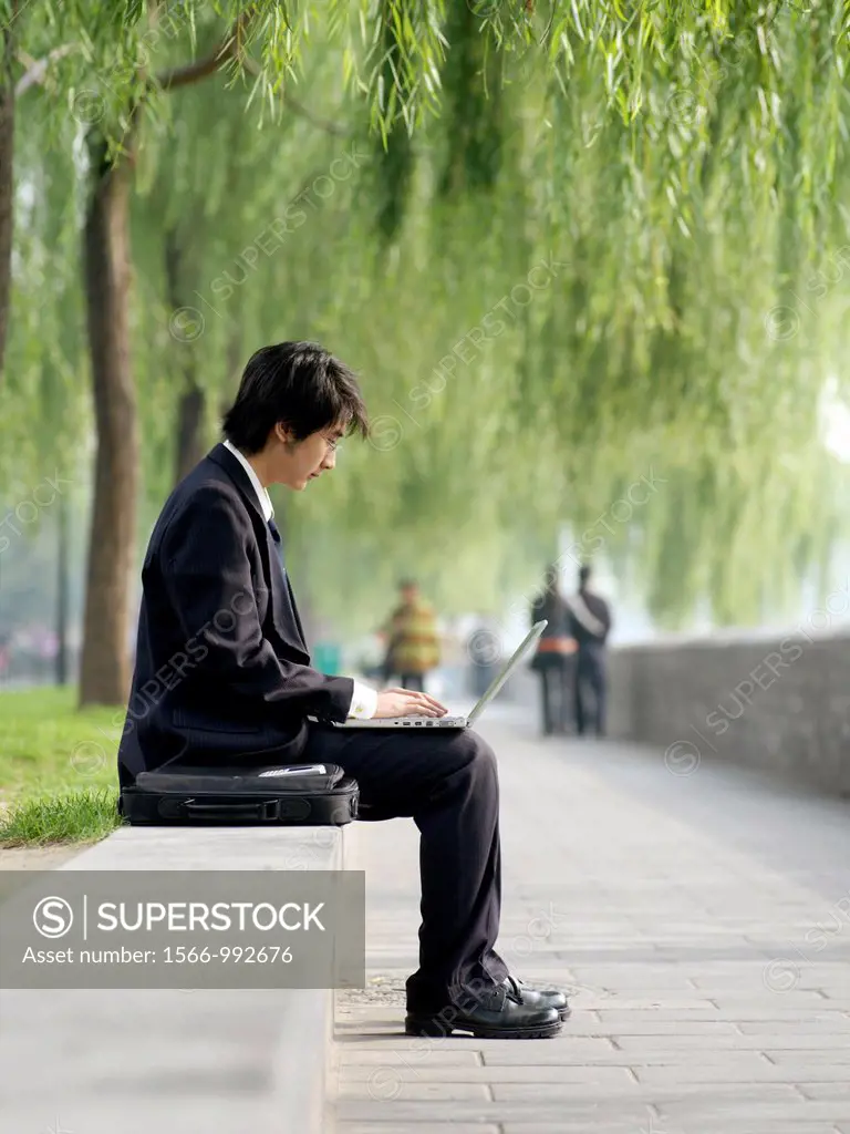A young Chinese business man in a suit checking his email on his laptop through wireless internet in a nature park