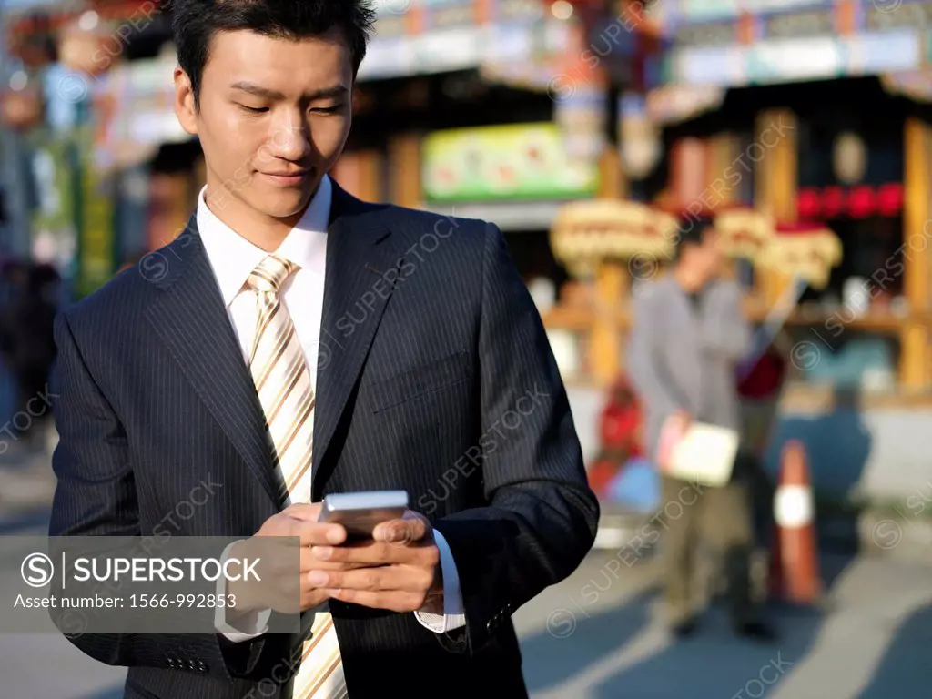 A young Chinese businessman in a suit checking his text messages or email on his mobile smart phone through wireless internet with traditional Chinese...
