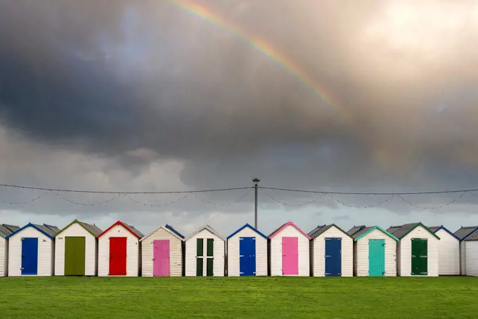 UK, England, Devonshire, Paignton. A rainbow in the sky above colourful beachside changing huts on the south coast of England.