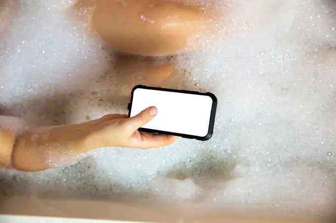 young female holding blank mobile phone, smart phone in bathtub with soapy foam close-up top view background copy space.