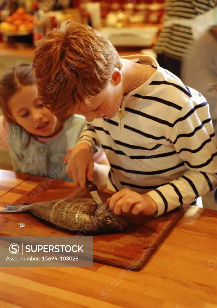 Two children in kitchen, one cutting a fish, parent in background