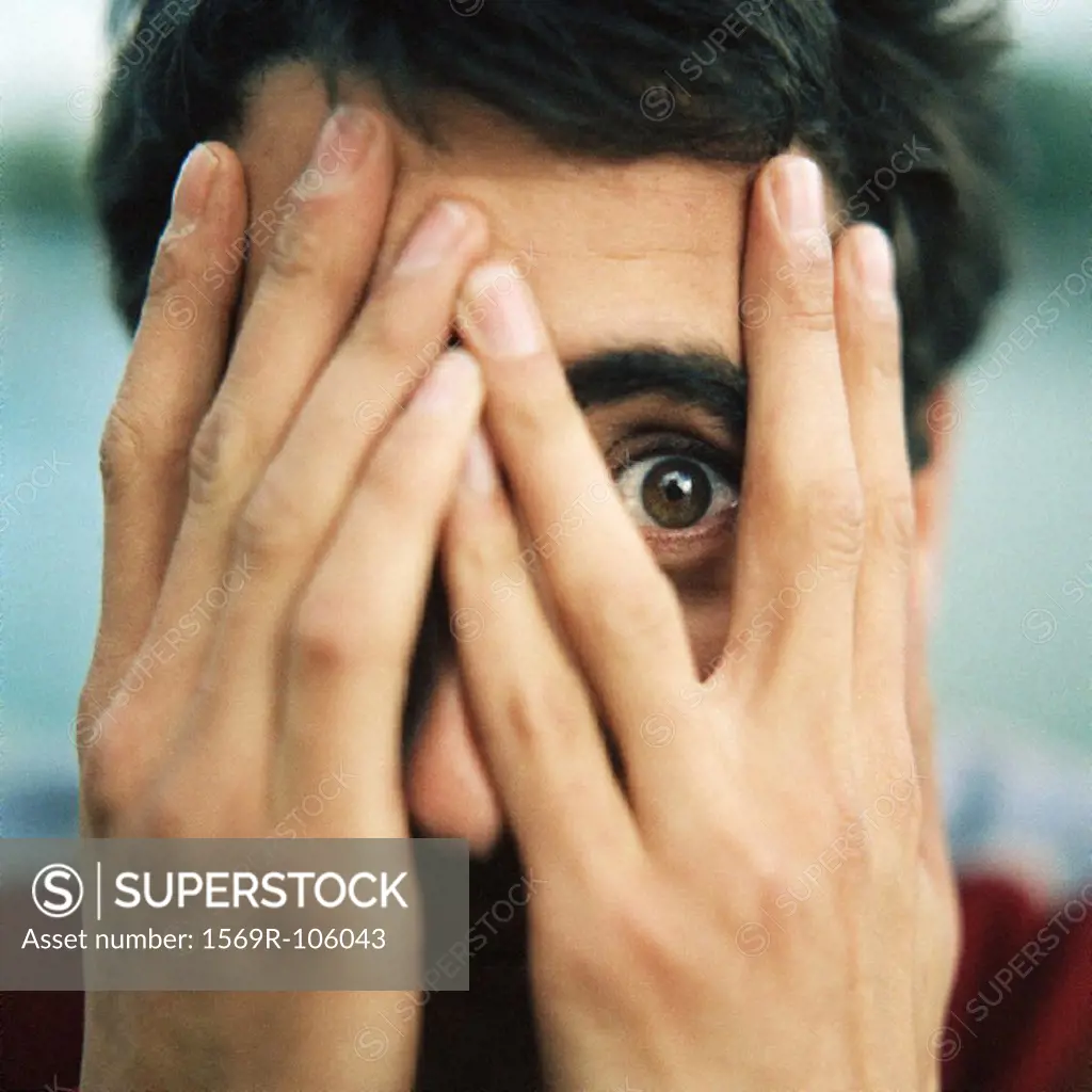 Young man with hands over face, looking through fingers with one eye, close-up