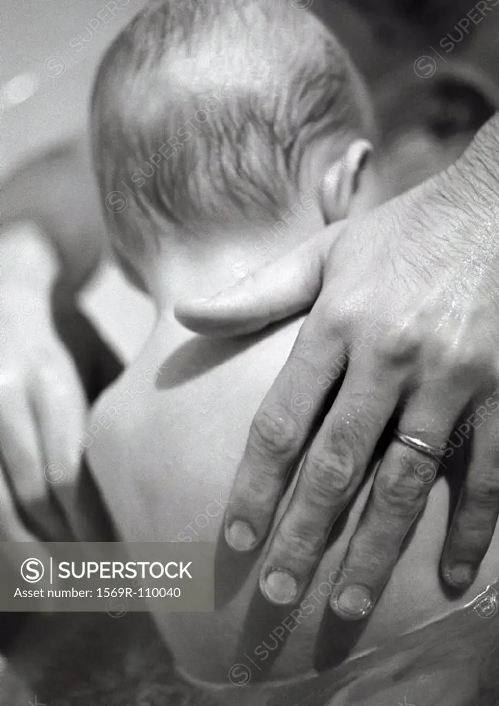 Baby sitting in bath, father´s hand on back, rear view, close-up, b&w