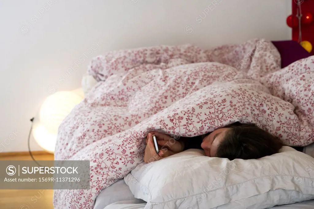 Woman lying in bed under covers, looking at smartphone