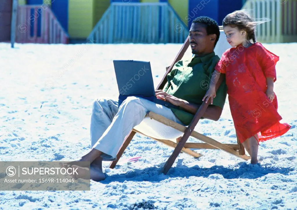 Man sitting in chair on beach using laptop, little girl standing by his side