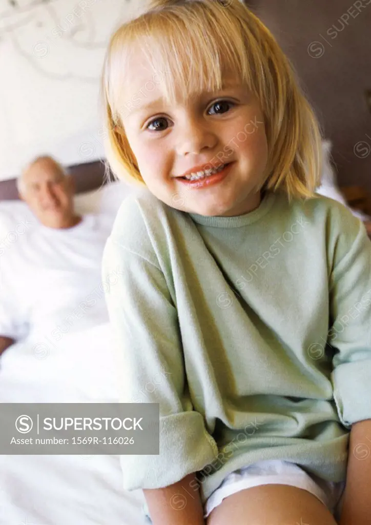 Small girl sitting on bed smiling, portrait, grandfather in background