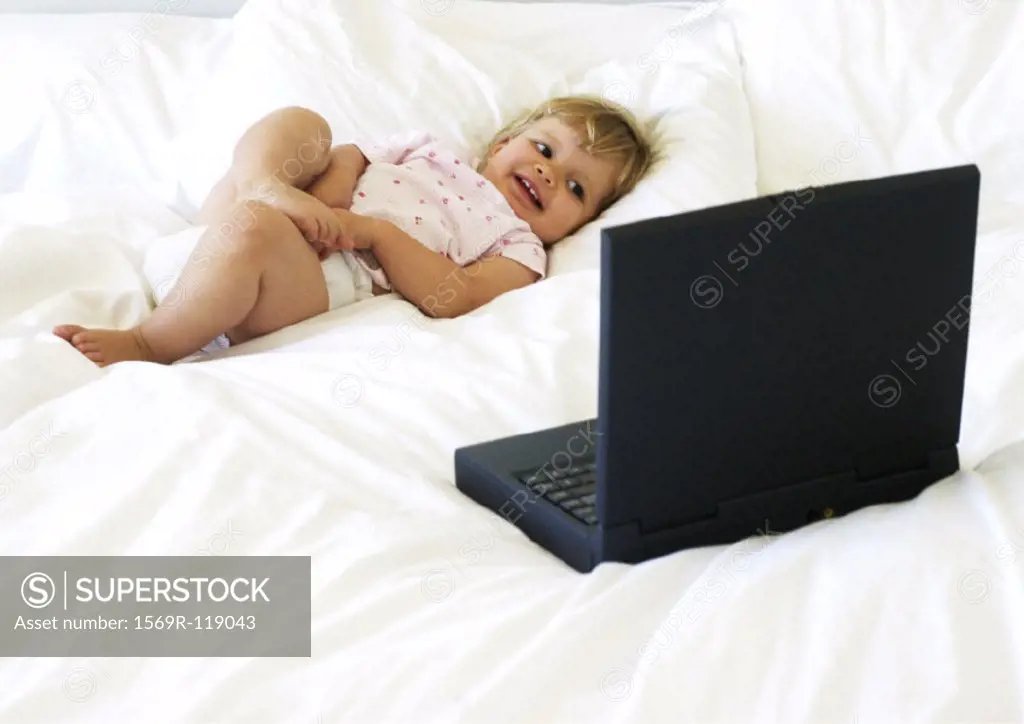 Baby lying on bed, looking at laptop computer