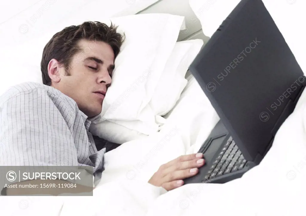 Man lying on bed with eyes closed, hand on laptop computer