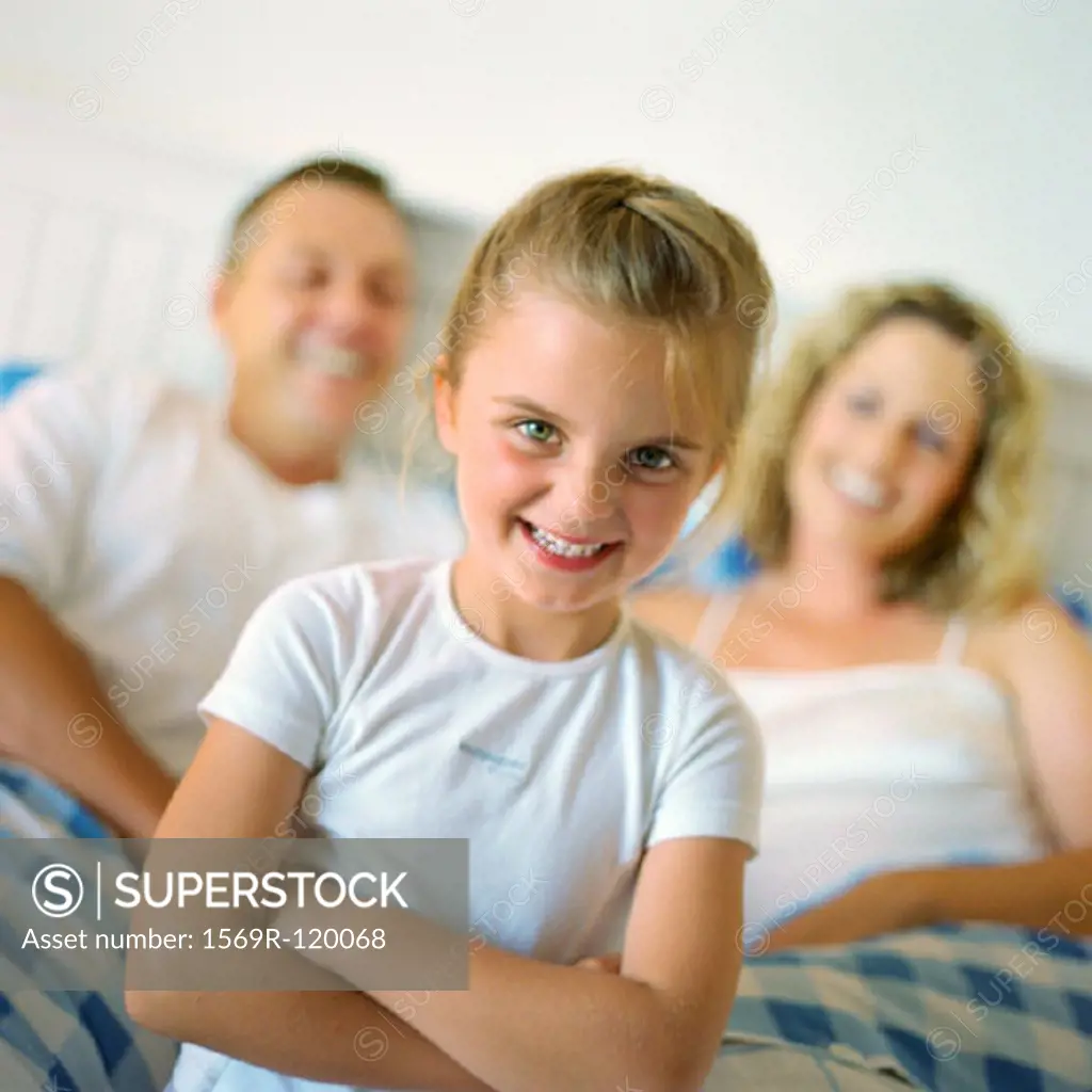 Girl folding arms and smiling in front of parents in bed, portrait