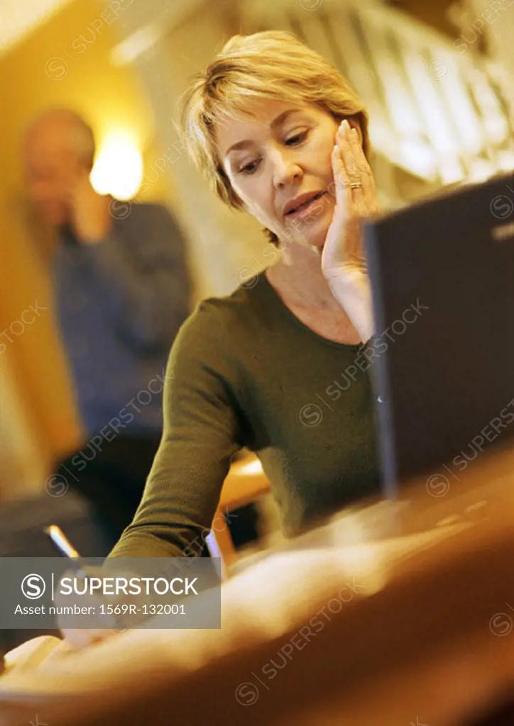 Mature woman using laptop, taking notes, man using cell phone in background