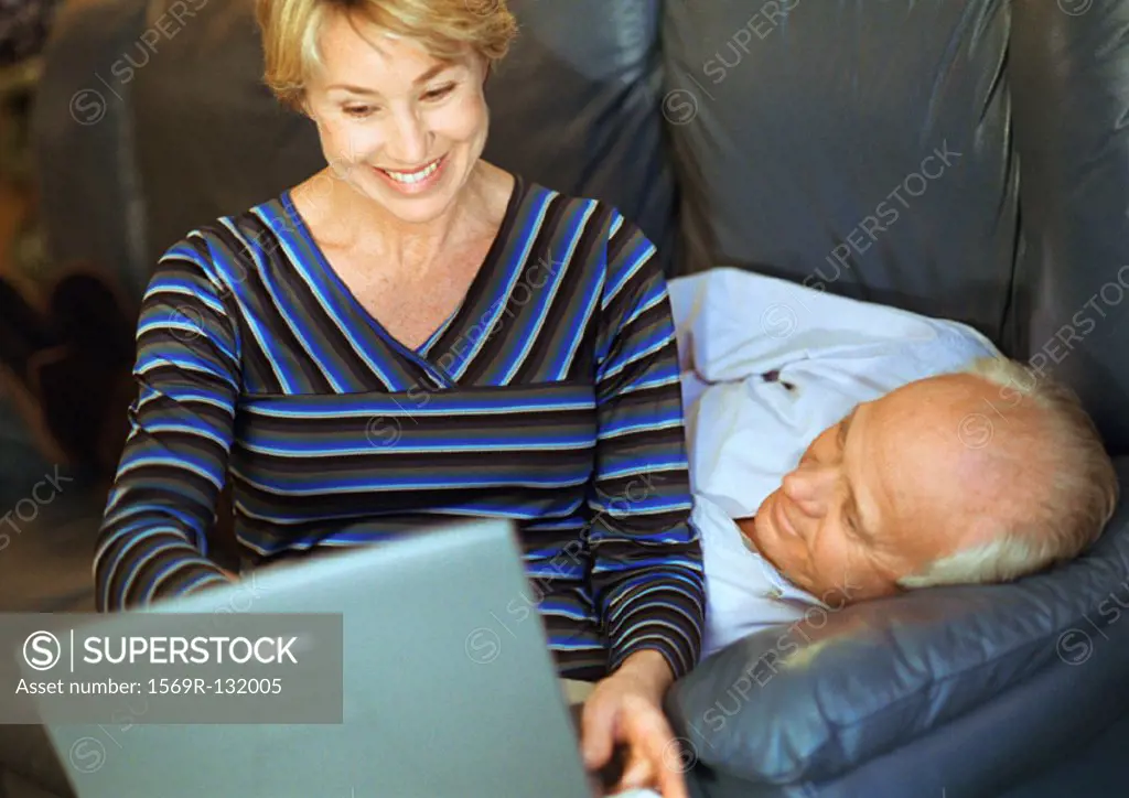 Mature couple, woman using laptop, man lying down
