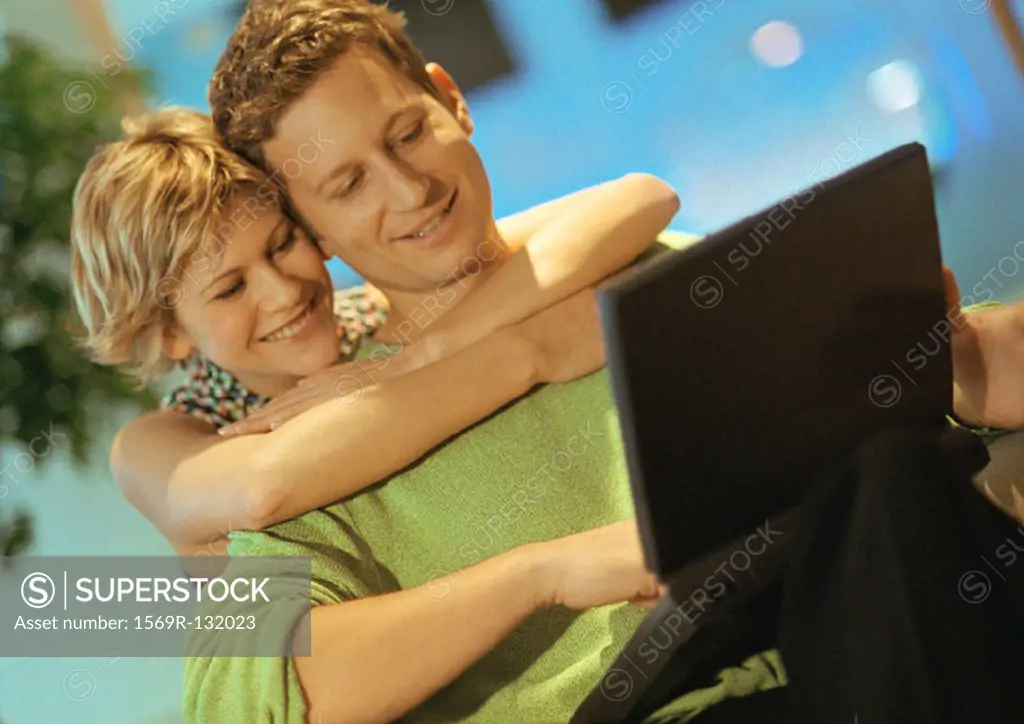 Couple smiling, man using laptop, woman with arms around man
