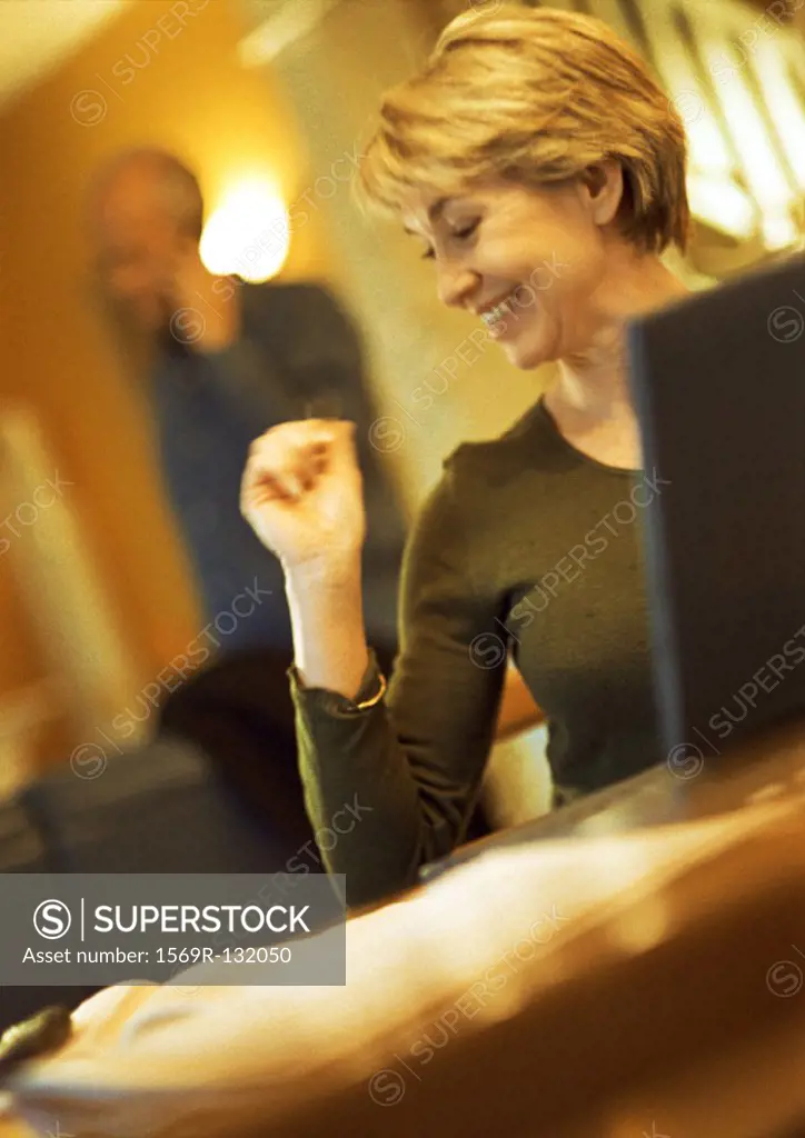 Mature woman smiling, using laptop, head turned, man using cell phone in background